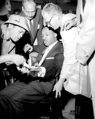 Dr. Martin Luther King, Jr. sits calmly with a letter opener protruding from his chest as Nettie C. Jackson tends his cut hand. September 20, 1958